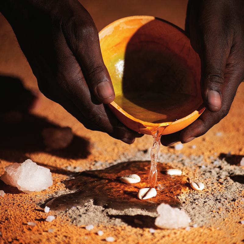 Two hands pour water from a calabash onto cowrie shells, salt and ash on red earth.